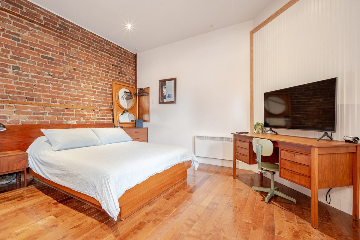 A spacious room features a comfortable bed with soft linens, positioned against a rustic brick wall. A wooden desk with a chair and a television are visible, complemented by subtle decorative accents. Natural light fills the space, highlighting the polished wooden floors.