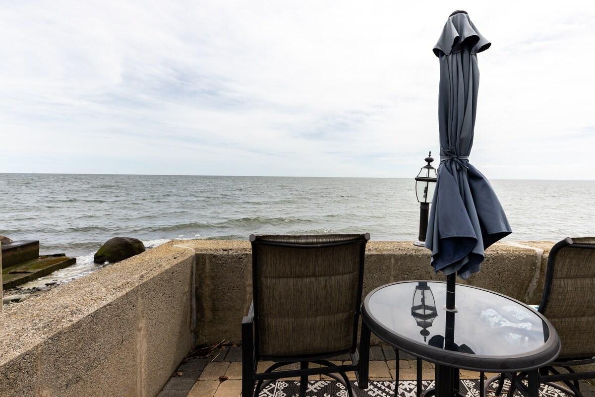 A bistro table with a glass top and two chairs is positioned by the sea wall, partially shaded by a dark blue umbrella. The expansive view of Long Island Sound stretches out beyond, with calm waters extending to the horizon under a light clouded sky.