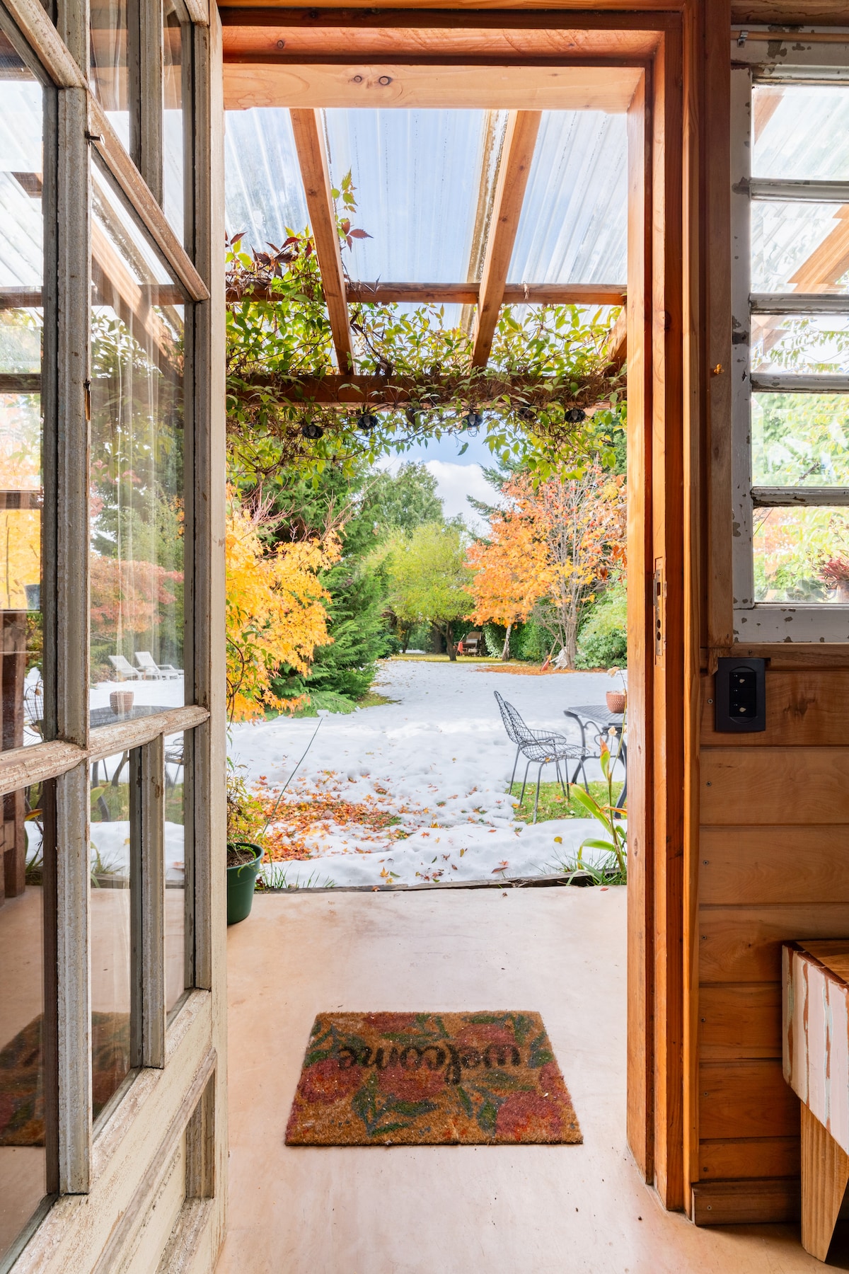 An inviting entrance is framed by wooden doors, leading to a view of vibrant autumn foliage and a private outdoor seating area. The welcome mat rests at the threshold, while natural light illuminates the interior space.