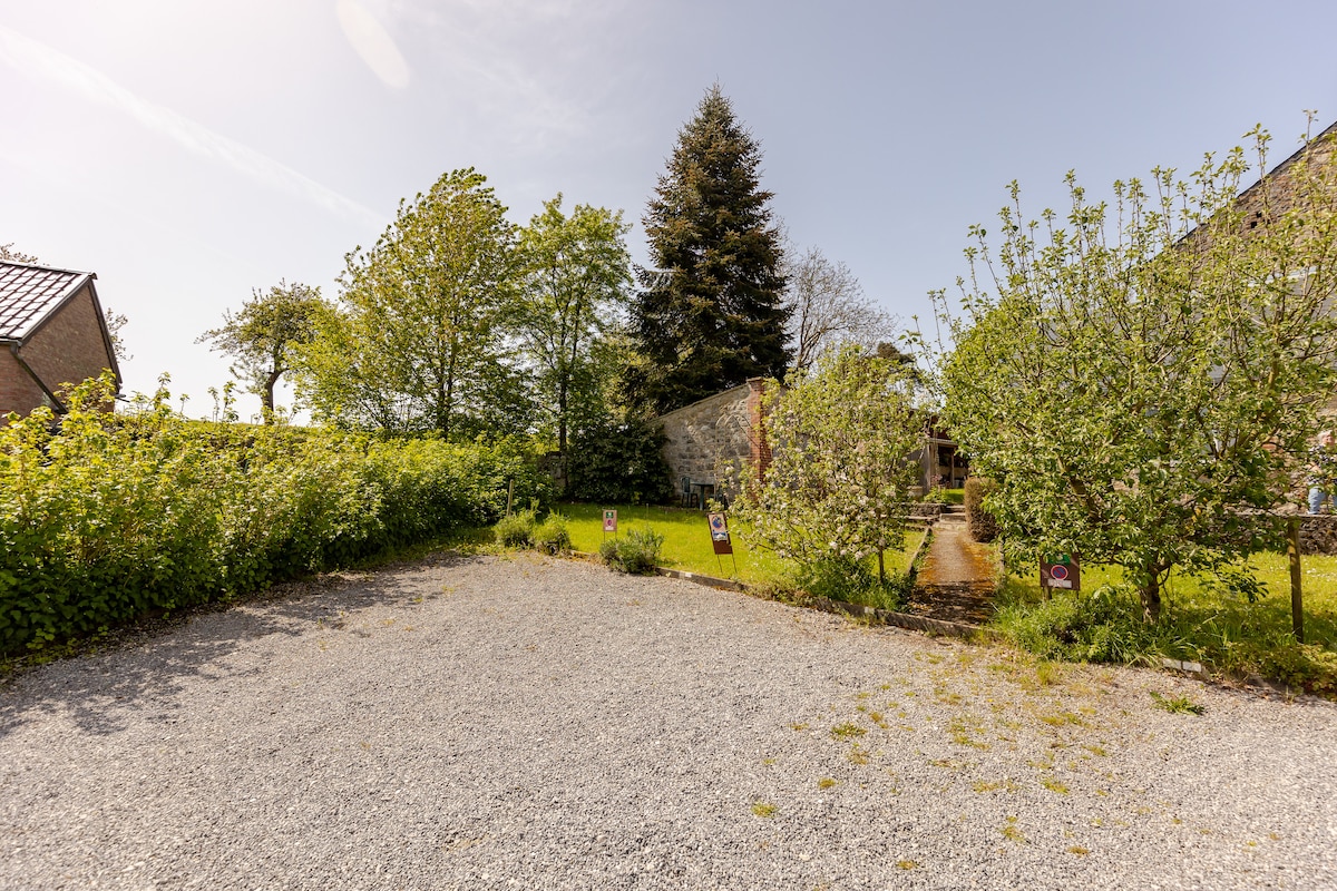 A spacious gravel parking area is surrounded by greenery, featuring low shrubs and trees. The entrance to the gîte is visible in the background, framed by plants. Natural sunlight brightens the scene, highlighting the tranquil rural setting.