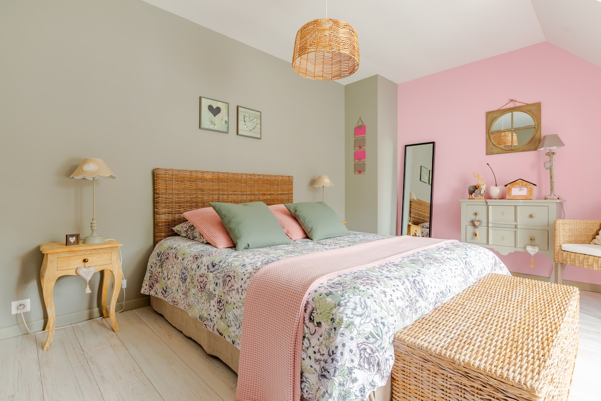 A spacious bedroom is featured, showcasing a woven headboard with decorative pillows. Soft pastel tones of pink and green accent the walls. A bedside table and lamp are present, along with a chest at the foot of the bed. Natural light enters through a nearby window.