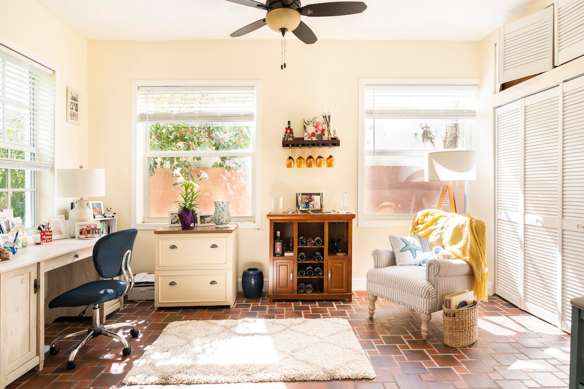 A brightly lit room features large windows allowing natural light to fill the space. A cozy chair with a yellow throw sits beside a low cabinet displaying decorative items. A desk with a rolling chair is positioned beside an area rug, enhancing the room's welcoming ambiance.