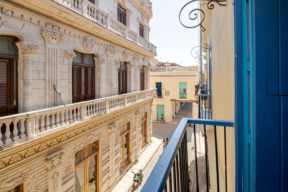 A view from the balcony showcases the charming architecture of colonial buildings, characterized by decorative balustrades and detailed façades. Soft sunlight highlights the narrow street below, while colorful doors invite exploration. The scene is framed by the balcony's iron railing, creating a cozy vantage point.
