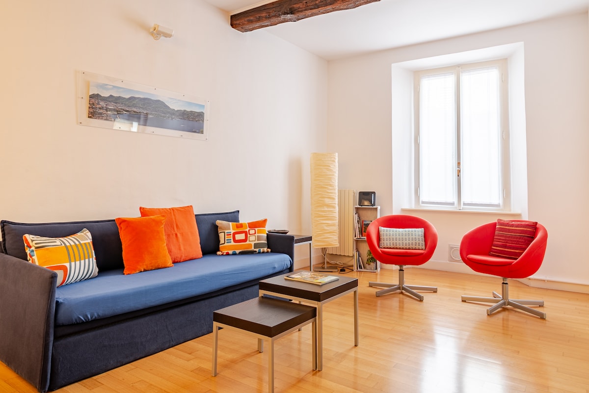 A modern living area is highlighted by a navy blue sofa adorned with colorful cushions in orange and yellow. A low coffee table stands in front, accompanied by two bright red chairs. Natural light enters through a window, accentuating the warm wooden floor.
