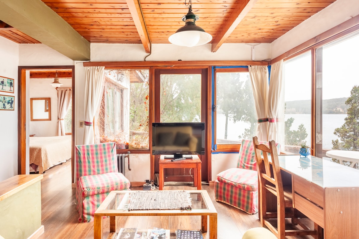 A bright, open living space features large windows that provide views of the lake. The area includes a television and a wooden table surrounded by chairs. Cozy seating is arranged using plaid-patterned cushions, enhancing the inviting atmosphere of the cabin.