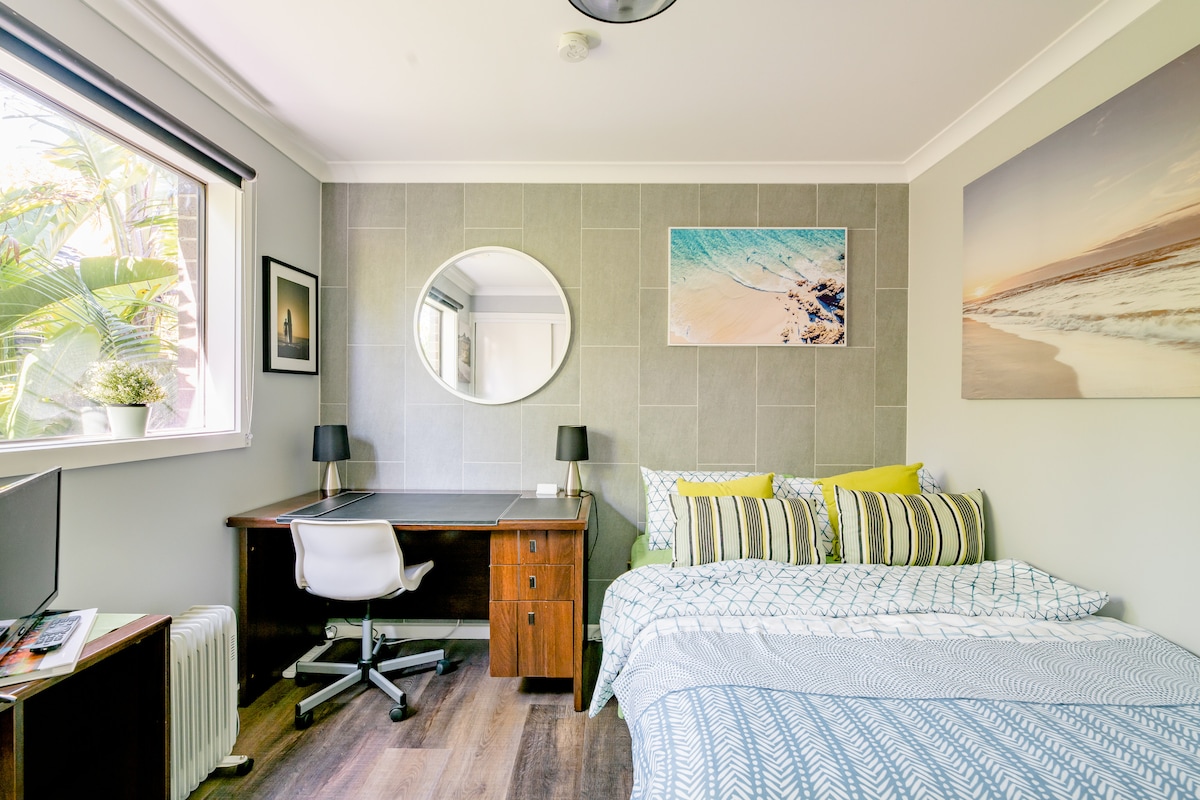 A cozy room features a spacious bed adorned with striped bedding and bright yellow accent pillows. A large window allows natural light to fill the space, highlighting a stylish desk with a chair and decorative artwork on the walls. A round mirror complements the decor.