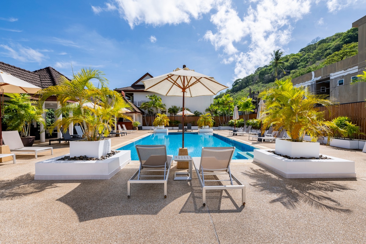 A large swimming pool is surrounded by lounge chairs under umbrellas. Tropical plants are positioned around the pool area, enhancing the serene atmosphere. The backdrop features a gently sloping hillside and various buildings, all under a clear blue sky.
