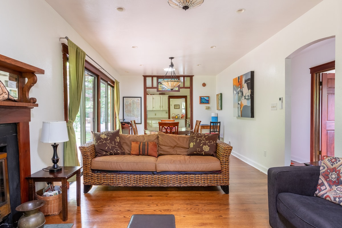 An open living area is highlighted with natural light from large windows. A woven couch with decorative pillows is positioned towards the center, accompanied by a dark armchair. The dining area is visible in the background, featuring a wooden table and colorful artwork on the walls.