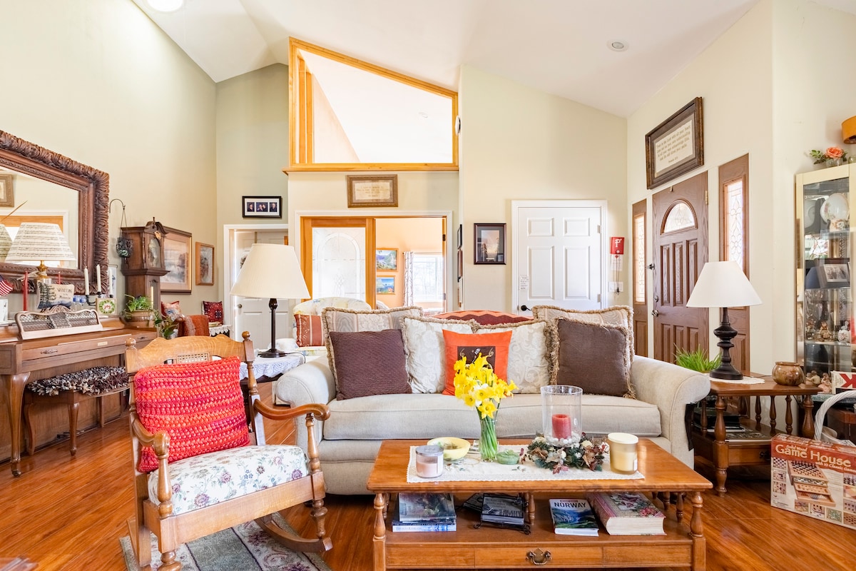 A warm living space is showcased with a spacious sofa accented by patterned cushions. A wooden coffee table sits in front, adorned with a candle and flowers. A vintage rocking chair is positioned nearby. Sunlight illuminates the room through large windows.