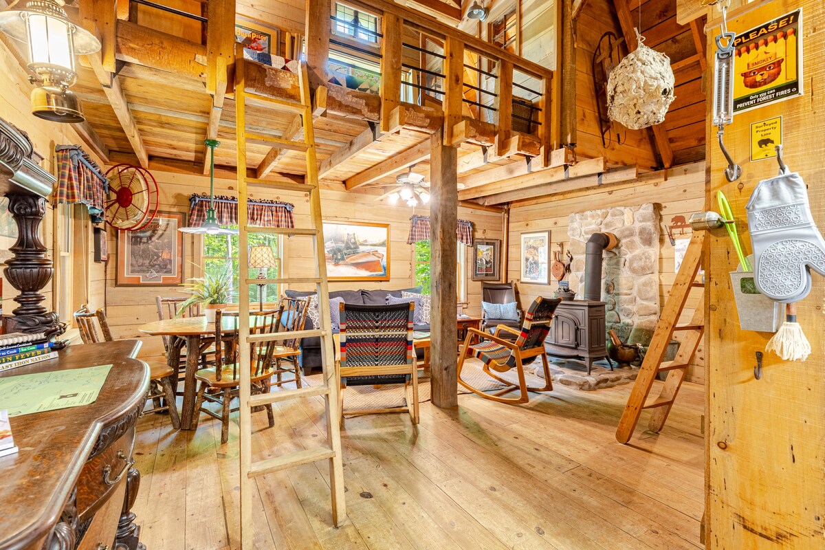 The interior of the cabin features warm wood paneling with exposed beams and a spacious layout. A dining area with a wooden table is positioned adjacent to a cozy seating area, which includes a wood-burning stove. A ladder leads to the lofted sleeping areas above.