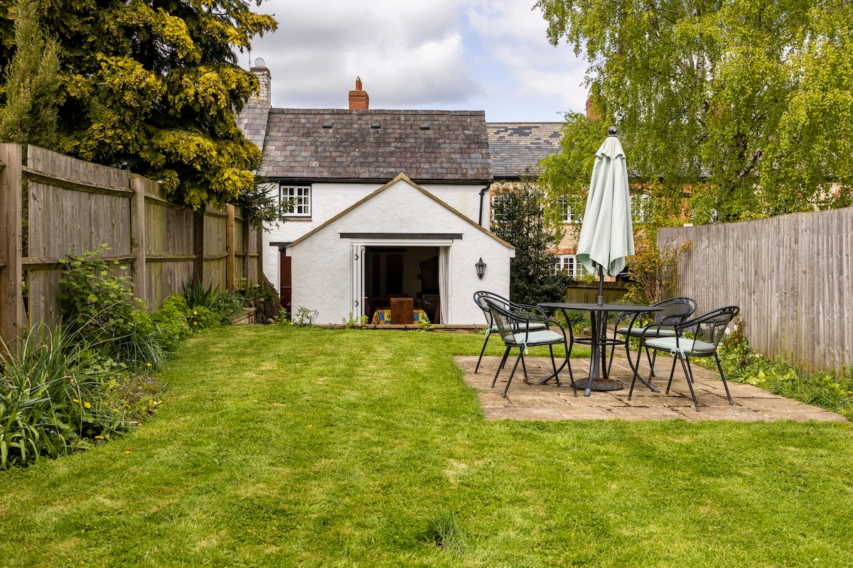 A grassy garden area is visible, bordered by a wooden fence. A patio with a round table and four chairs is positioned to the right, while a sunshade stands nearby. The white cottage is seen in the background, framed by greenery and under a partly cloudy sky.