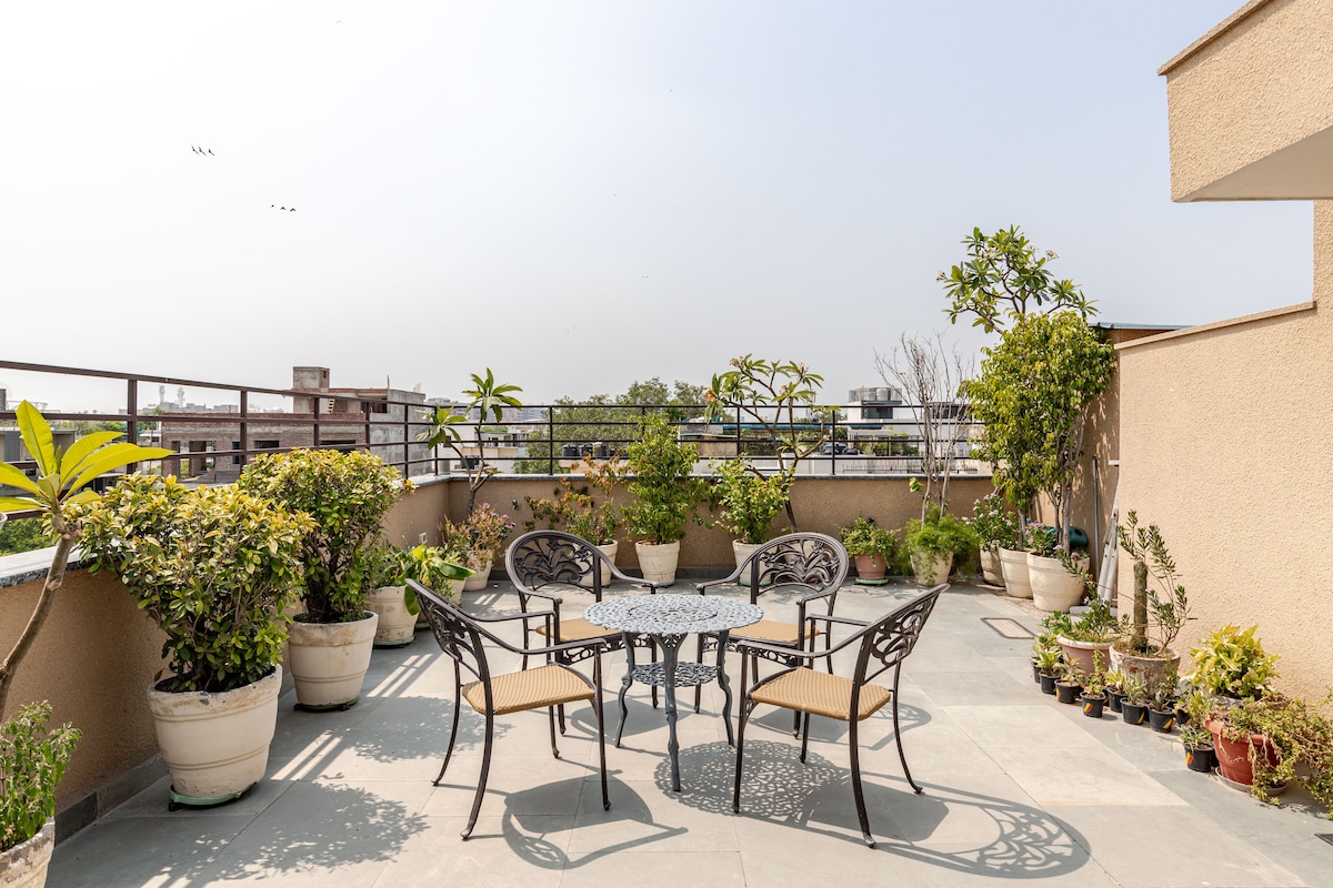 A spacious terrace is adorned with potted plants and small trees, creating a verdant oasis. Four elegant chairs surround a round table, inviting relaxation and outdoor gatherings. Clear skies above provide ample natural light to the inviting outdoor space.
