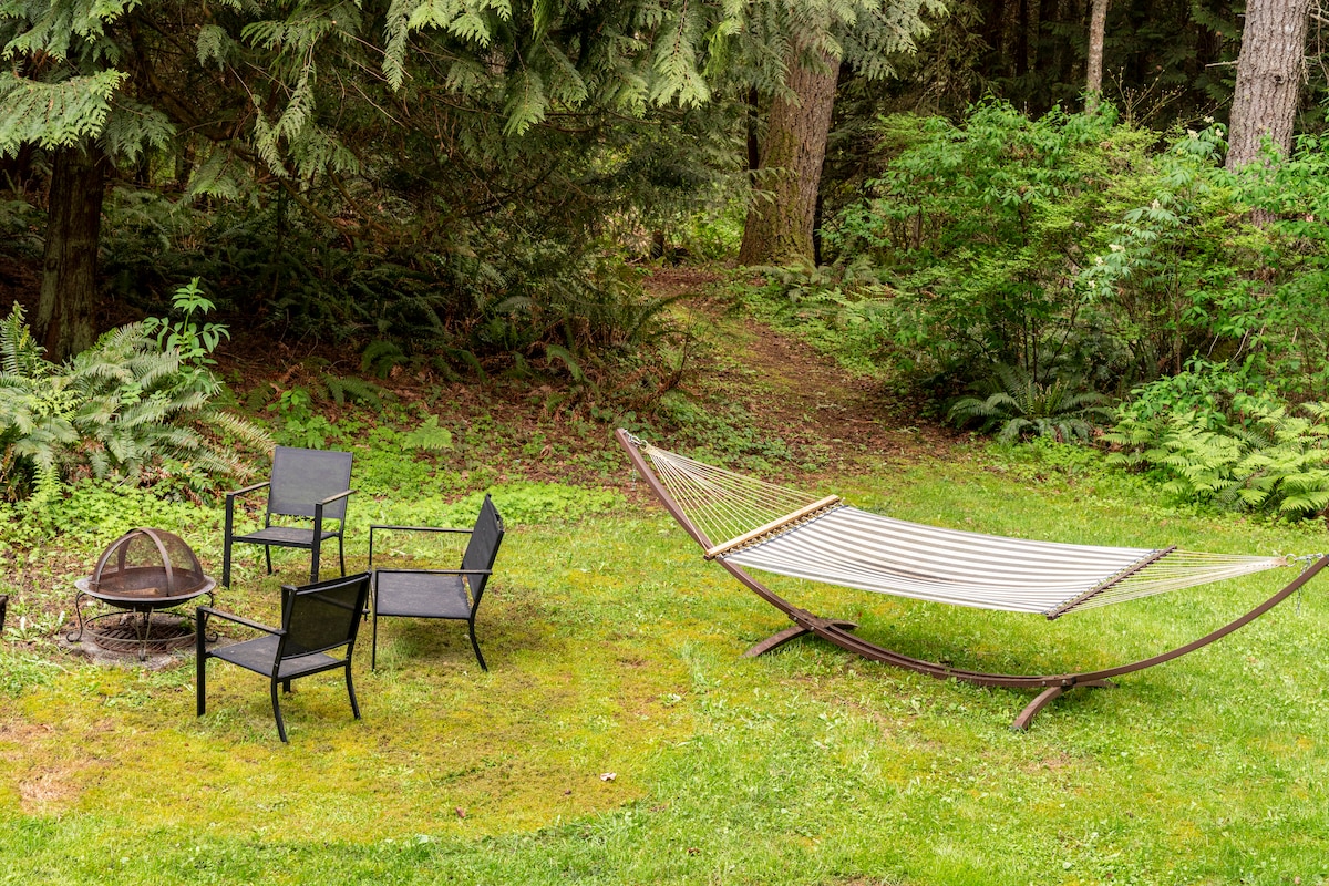A serene outdoor space features a hammock and a fire pit surrounded by four black chairs. Lush greenery and trees provide a natural backdrop, creating a tranquil setting ideal for relaxation.