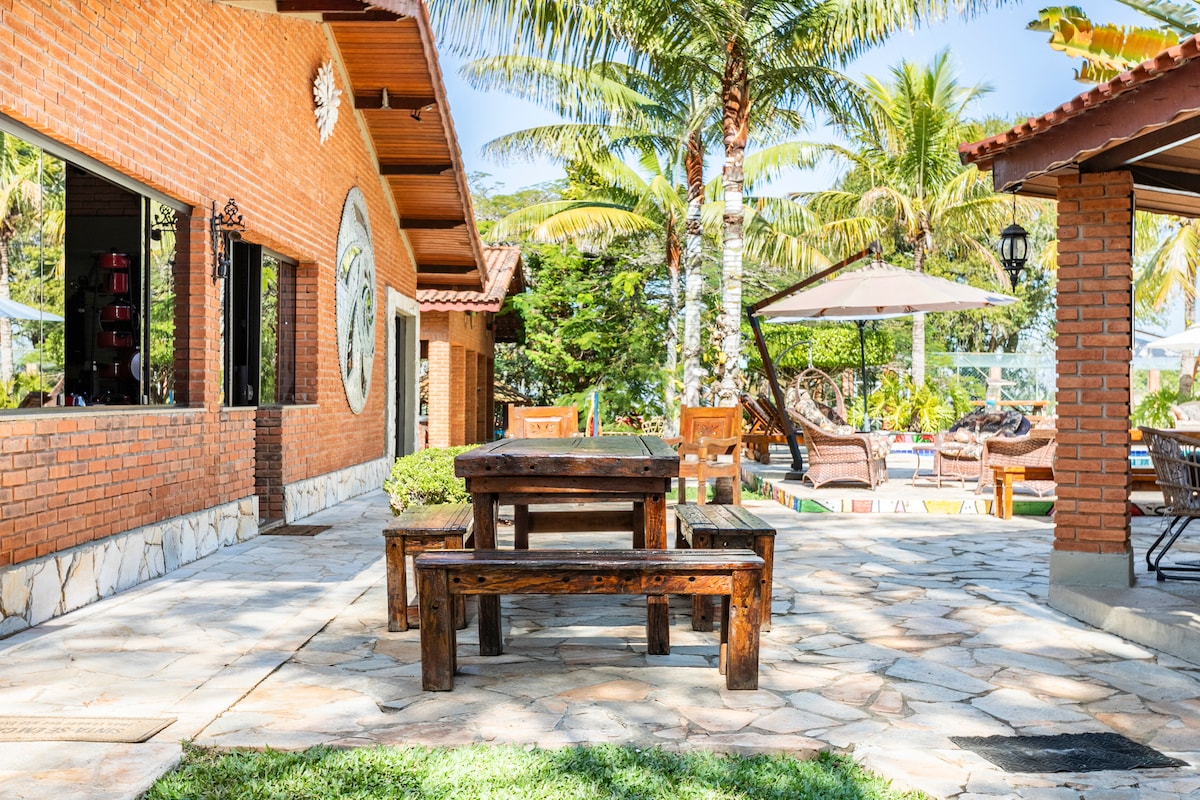 An outdoor dining area is situated on a stone patio, featuring a wooden table surrounded by chairs. Palms and lush greenery create a natural backdrop, with a glimpse of a pool and lounge area under a shaded gazebo in the distance.