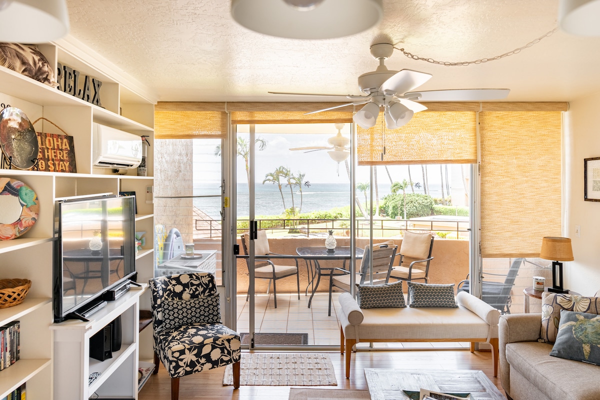 A light-filled living area is depicted with a cozy seating arrangement, including a sofa and patterned chair. Sliding glass doors open to a patio overlooking the ocean. Shelves display books and a television is positioned against a wall, adding to the welcoming ambiance.
