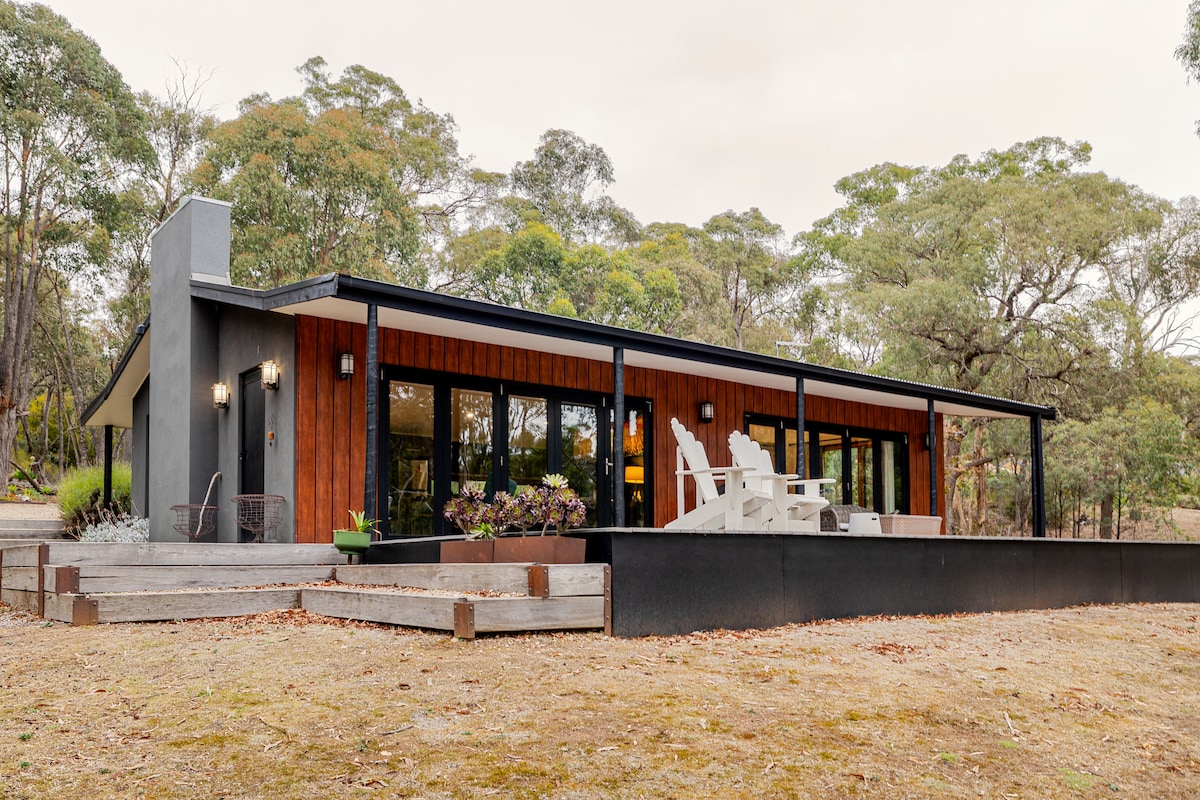 A modern retreat is showcased with a sleek exterior, featuring large glass doors that allow natural light to fill the interior. The wraparound deck offers ample outdoor space with stylish seating and views of the surrounding landscape.