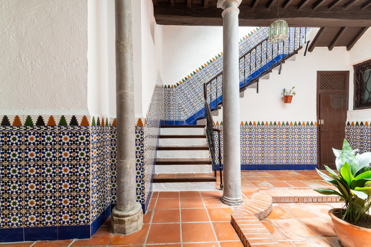 An elegant entryway is adorned with traditional tile work, featuring intricate patterns in blue, white, and earthy tones. Wrought iron railings outline the staircase, which is complemented by wooden steps. A column supports the ceiling, and natural light illuminates the space.