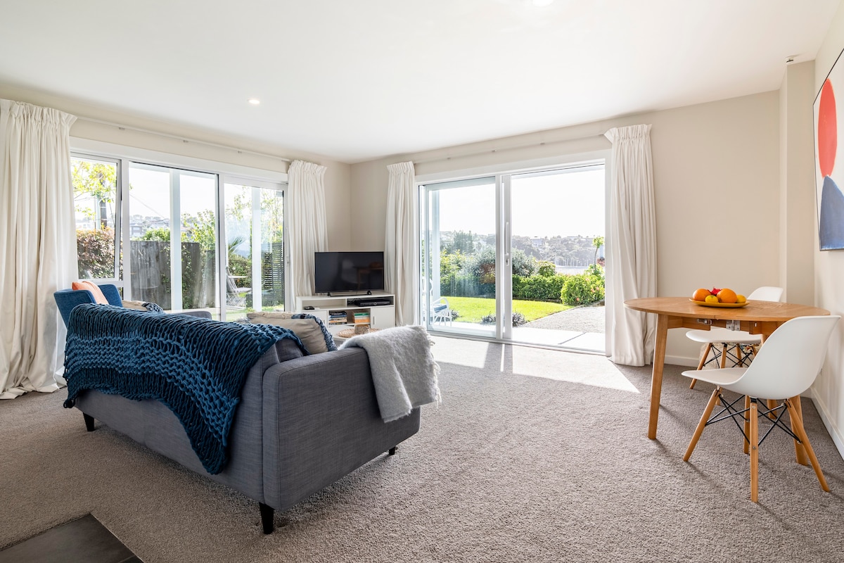A spacious living area features large glass doors that open to a private outdoor patio, allowing natural light to fill the room. A comfortable sofa with a textured throw is positioned near a television and a dining table with fruit. Soft, neutral tones enhance the inviting space.