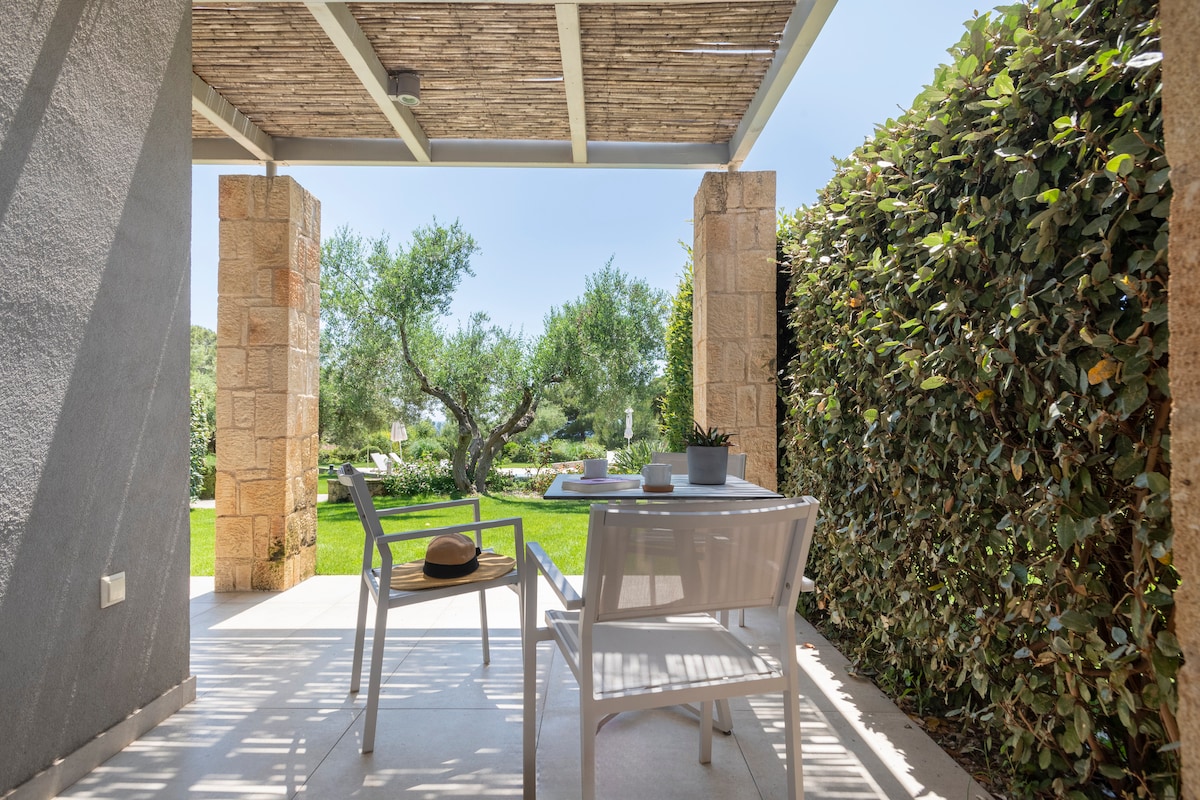 A shaded outdoor seating area features a small table and two chairs, offering views of a lush green garden with olive trees. The surrounding hedges create a sense of privacy, enhancing the tranquil atmosphere of the space.