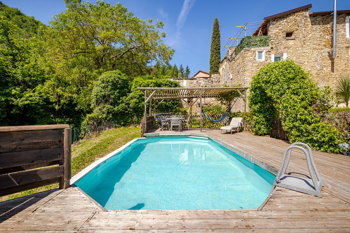 A serene pool area is surrounded by greenery, featuring a private pool with a wooden deck. Lounge chairs are positioned nearby, and a shaded dining space under a pergola is visible. The backdrop includes stone buildings that reflect the rich history of the hamlet.