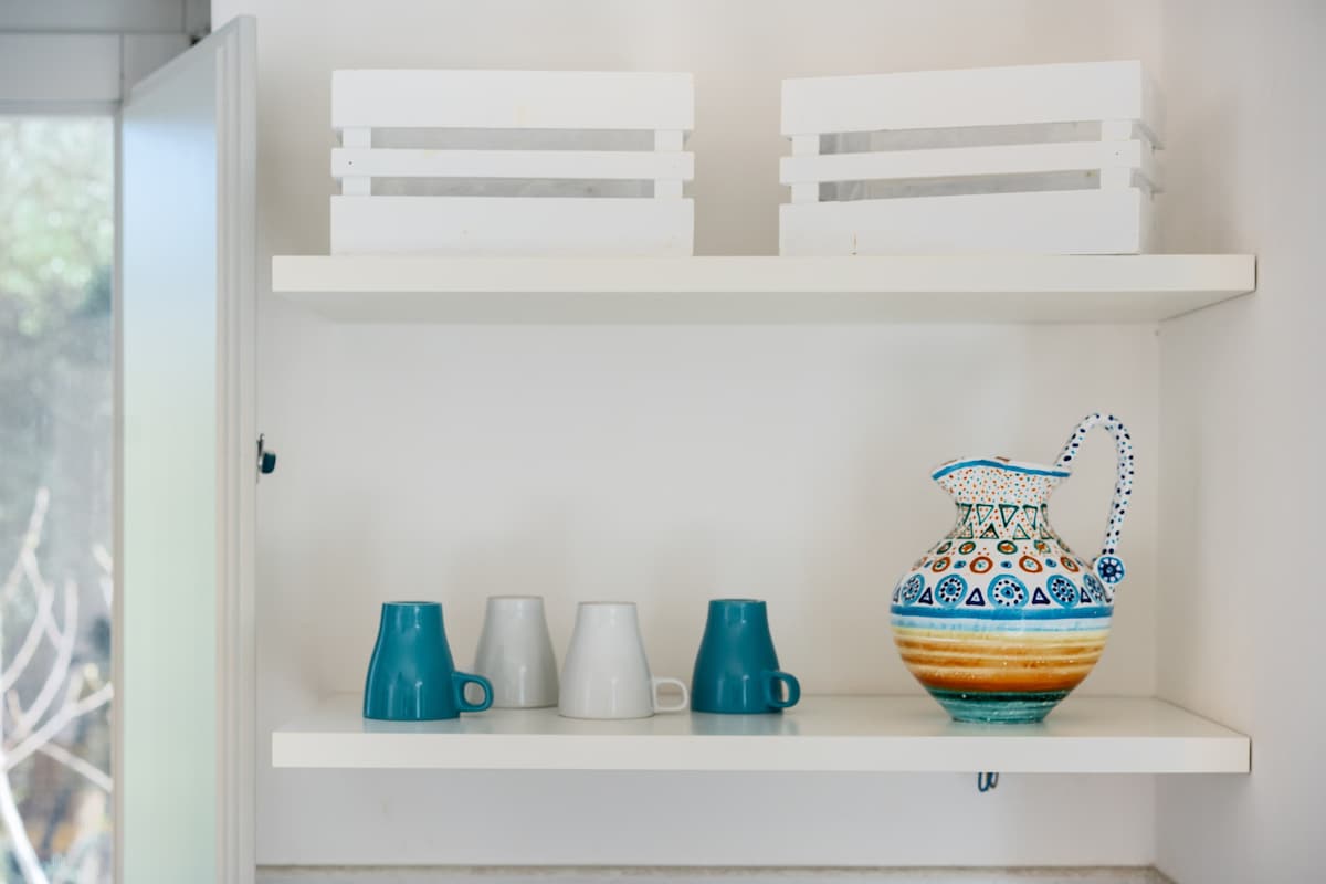 A kitchenette shelf is displayed, featuring a colorful ceramic pitcher alongside six cups in varying shades of blue and gray. Two white storage boxes are positioned above, contributing to the organized appearance of the space.