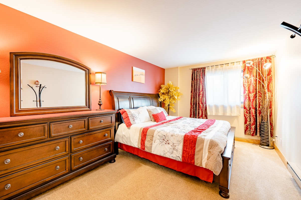 A private bedroom features a traditional wooden dresser and a large mirror. The bed is dressed with a floral-patterned blanket and red accents, complemented by decorative curtains. Natural light enters through the window, highlighting the warm tones of the walls and the room's spacious layout.