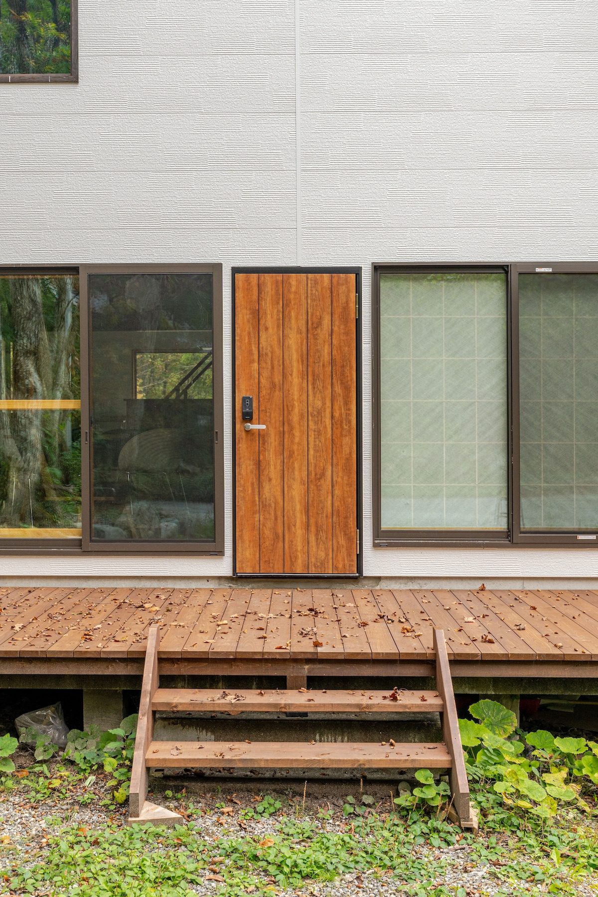 The cottage entrance is framed by large windows on either side, allowing natural light to fill the space. A wooden door with a sleek design serves as the focal point, complemented by a small set of steps leading up to a cozy wooden deck surrounded by foliage.