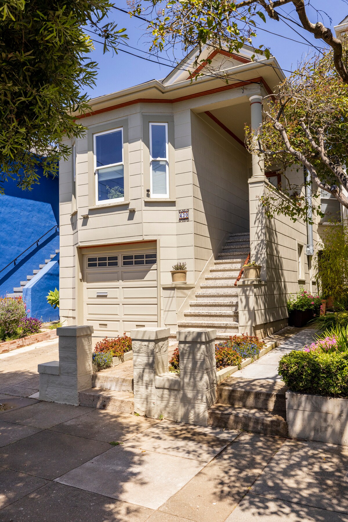 A charming exterior of a two-story home is framed by well-maintained gardens. The entrance features a set of stairs leading to a private entrance, flanked by small potted plants. Large windows provide natural light, and a garage is visible at the base of the structure.