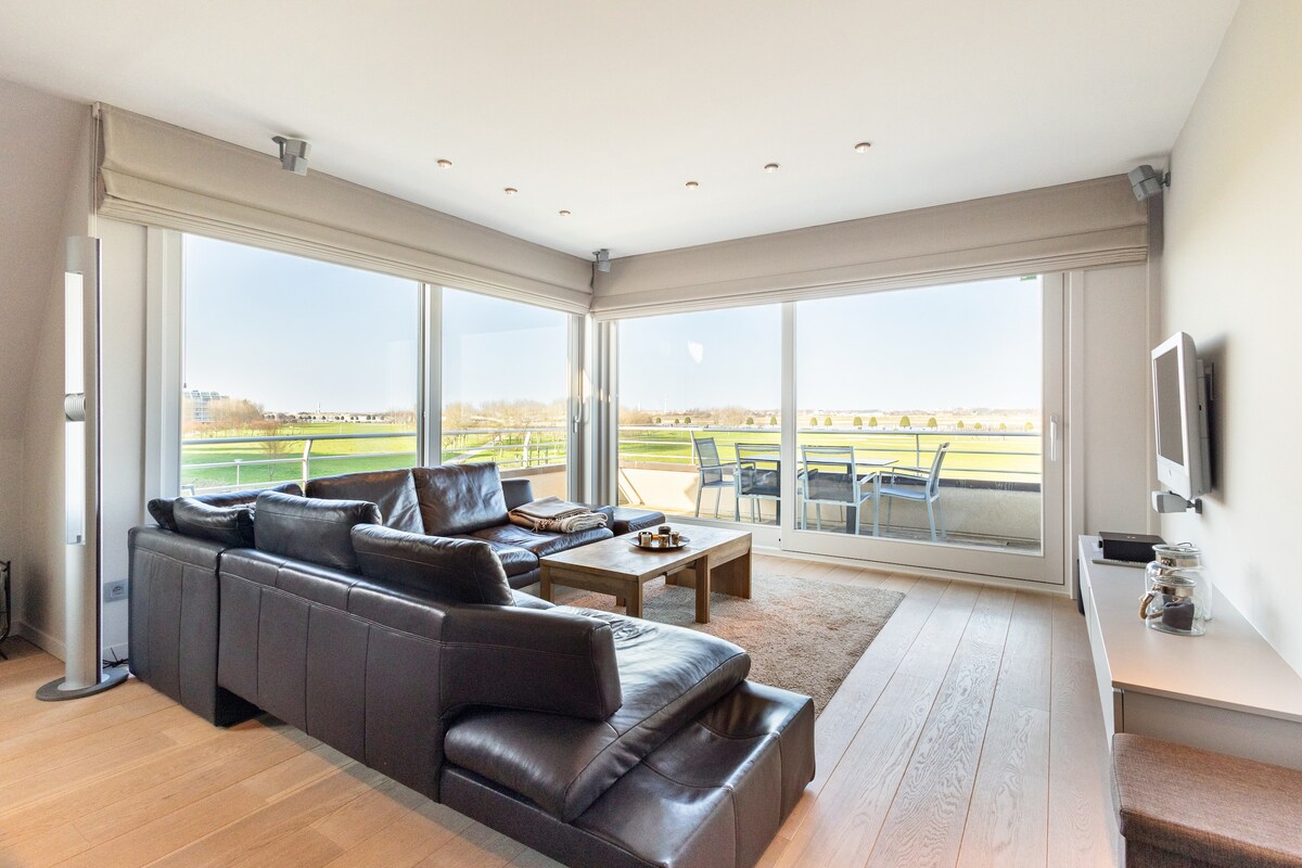 A spacious living area features a large sectional sofa and a wooden coffee table. Expansive glass doors provide a view of the outdoor terrace and surrounding green landscape. Natural light fills the room, enhancing the calm ambiance.