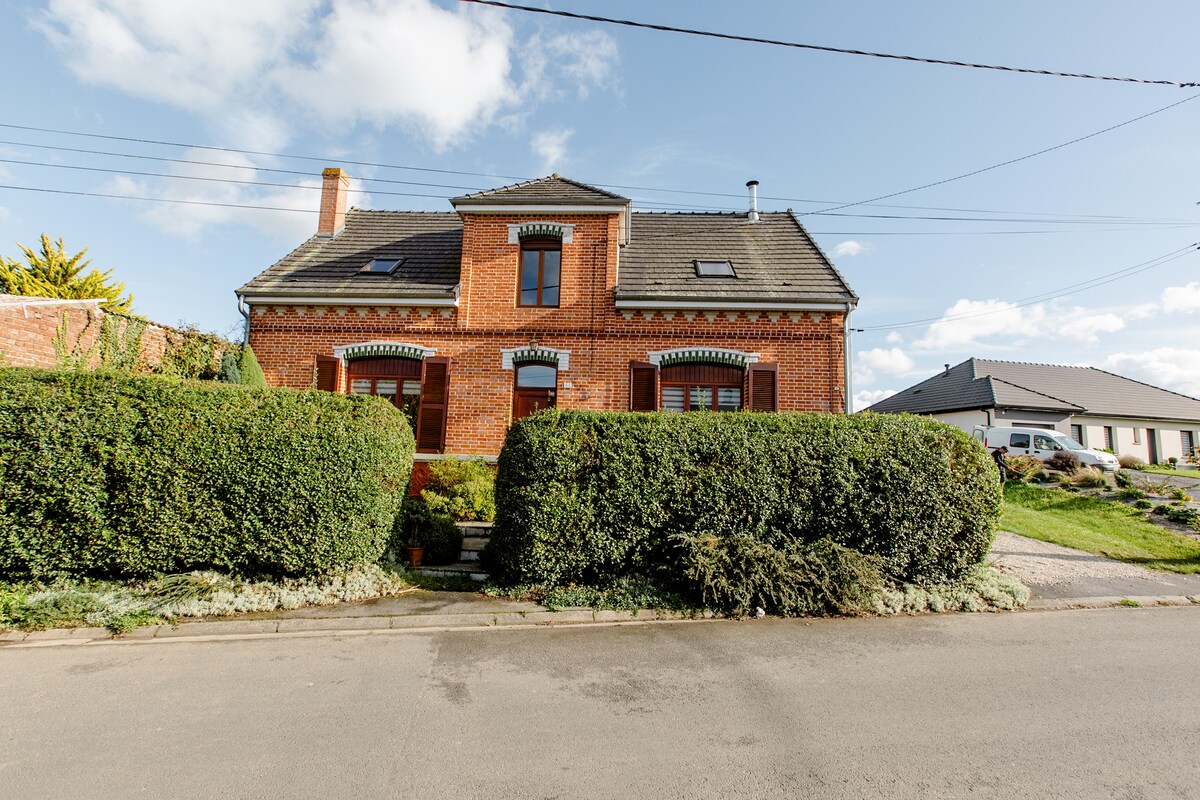 A charming brick house is surrounded by neatly trimmed hedges, creating a welcoming entrance. The structure features multiple windows and rooftops with chimneys, set against a clear blue sky. The front pathway leads to the main entrance, enhancing the overall inviting appeal.