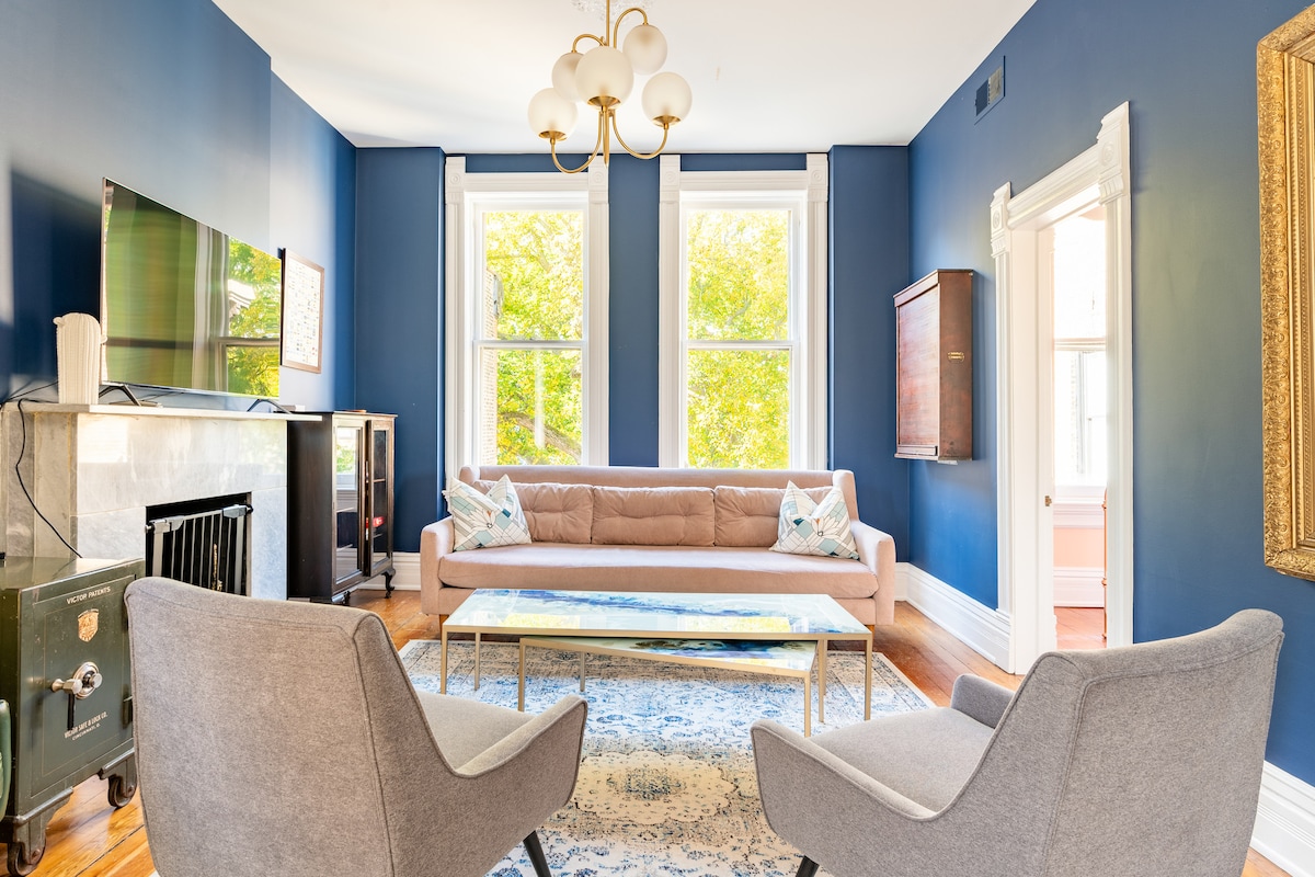 A bright living area features large windows allowing natural light to illuminate the space. A soft sofa is centered on a decorative area rug, complemented by two modern armchairs. A vintage safe and elegant chandelier enhance the unique character of the room.