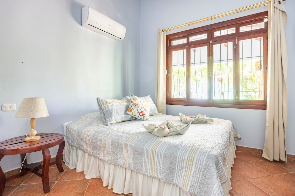A bright bedroom is equipped with air conditioning and a spacious bed adorned with decorative pillows. Natural light filters through a window secured with bars, while a small bedside table holds a lamp. The room features a cool blue wall that adds to its serene vibe.