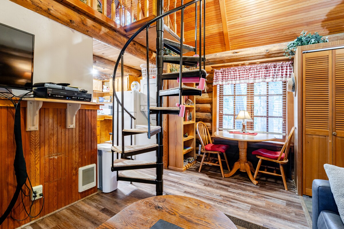 A spiral staircase leads to an upper level, surrounded by log walls and large windows that allow natural light to fill the space. A dining nook with a small table and red chairs is visible, along with a seating area featuring a dark couch and wall-mounted television.