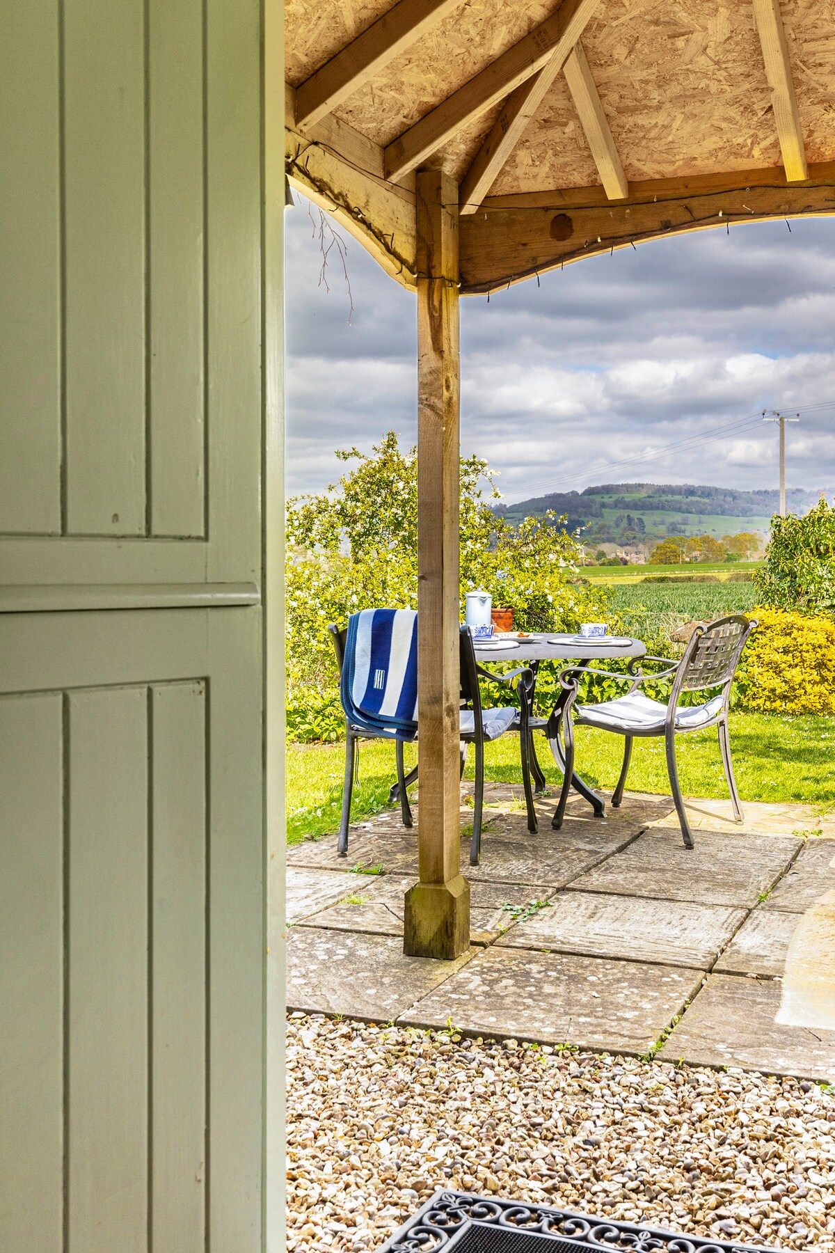 A view from the doorway reveals a small outdoor seating area with a table and four chairs under a roofed porch. Scenic rolling hills are visible in the background, framed by greenery and flowering shrubs. A pathway leads towards the sitting space.