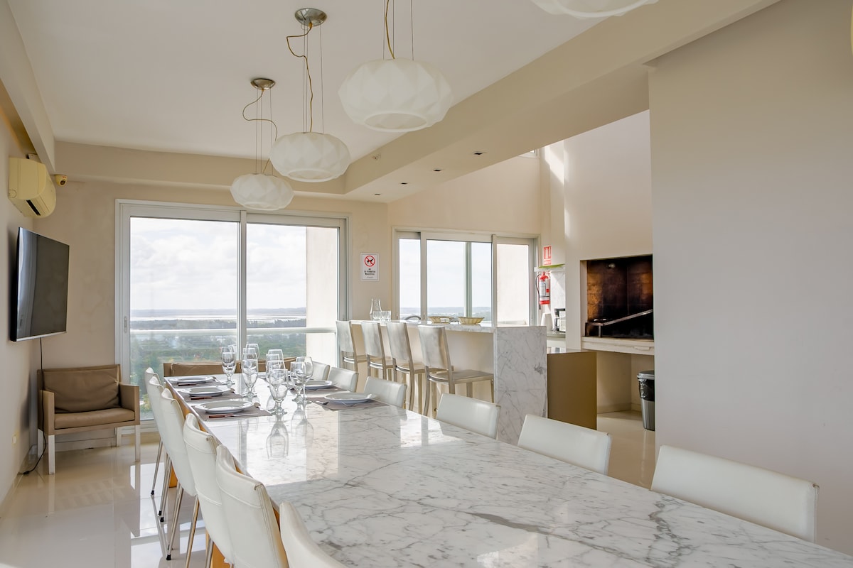 A stylish dining area features a large marble table with comfortable seating for multiple guests. Elegant pendant lights hang from the ceiling, illuminating the space. Floor-to-ceiling windows offer panoramic views of the surroundings, enhancing the overall airy ambiance.