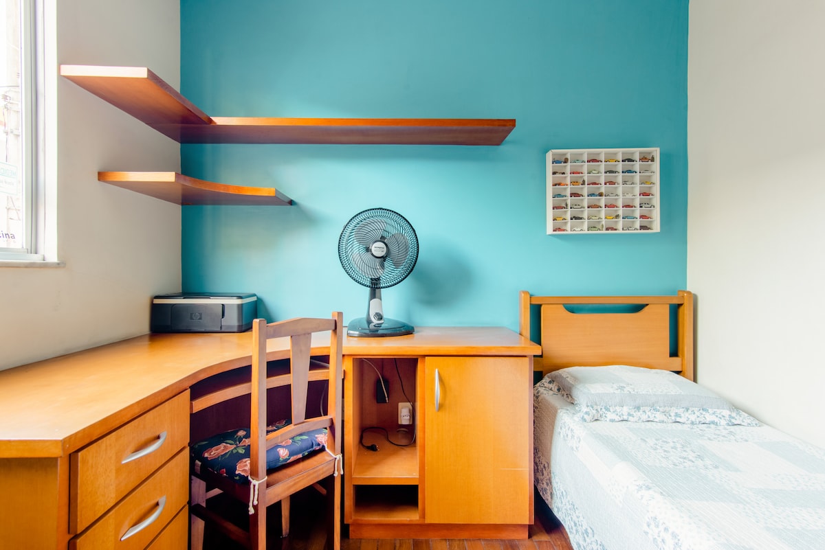 A calm bedroom features a single bed with light bedding against a blue wall. A wooden desk with a fan and a printer occupies one corner, complemented by shelves that hold various items. Natural light streams in from a nearby window.
