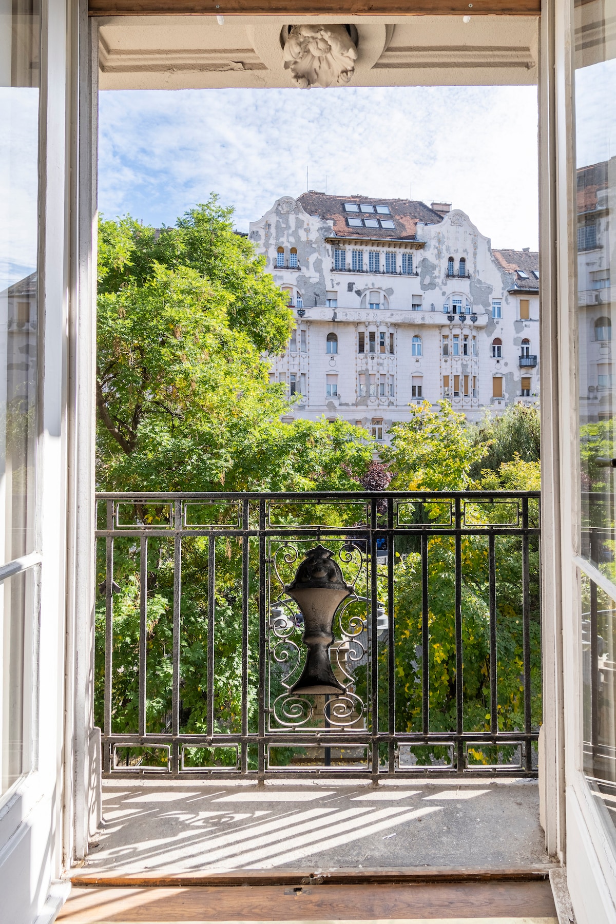 A view from a french balcony frames lush greenery and historical buildings in the background. The balustrade design offers a glimpse of the peaceful surroundings, with sunlight filtering through the leaves and illuminating the space.