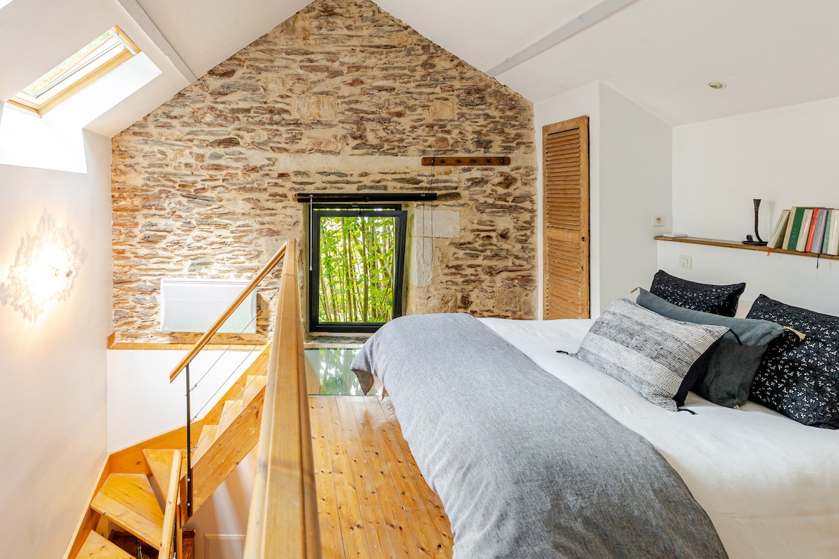 An inviting loft bedroom features textured stone walls and a sloped ceiling. A spacious bed is adorned with stylish black and white pillows, positioned near a window that frames views of greenery. A wooden staircase leads to the lower level, enhancing the room's open design.