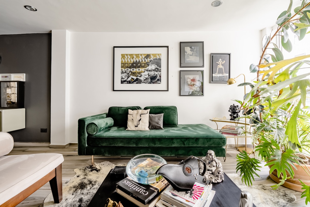 A modern living area features a green velvet sofa with decorative cushions, positioned against a white wall adorned with framed artwork. A large plant adds a touch of greenery, while a coffee table displays books and a decorative item, creating a stylish and inviting space.