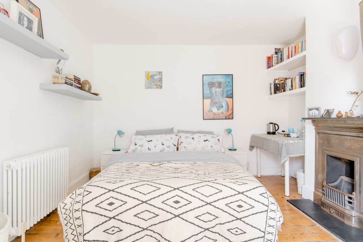 A bright bedroom is displayed, featuring a spacious bed with patterned bedding. Shelves are mounted on the white walls, adorned with books and decorative items. A small table with a kettle is positioned near a classic fireplace, contributing to a comfortable ambiance.