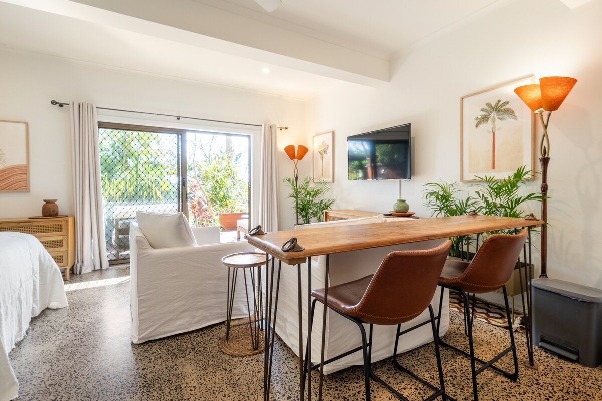 A bright and airy studio space features a wooden dining table with two leather chairs, positioned near a window. A cozy seating area includes a light-colored sofa, and a television is mounted on the wall. Potted plants add a touch of greenery.