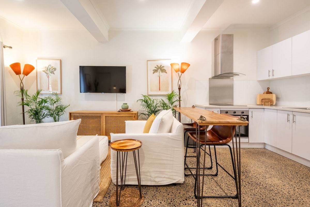 A bright living area features a comfortable white sofa and armchair, accentuated by warm floor lamps. A wooden dining table with high stools sits nearby. The kitchen is equipped with modern appliances, and tropical-themed artwork adorns the walls alongside potted plants, bringing a fresh touch to the space.