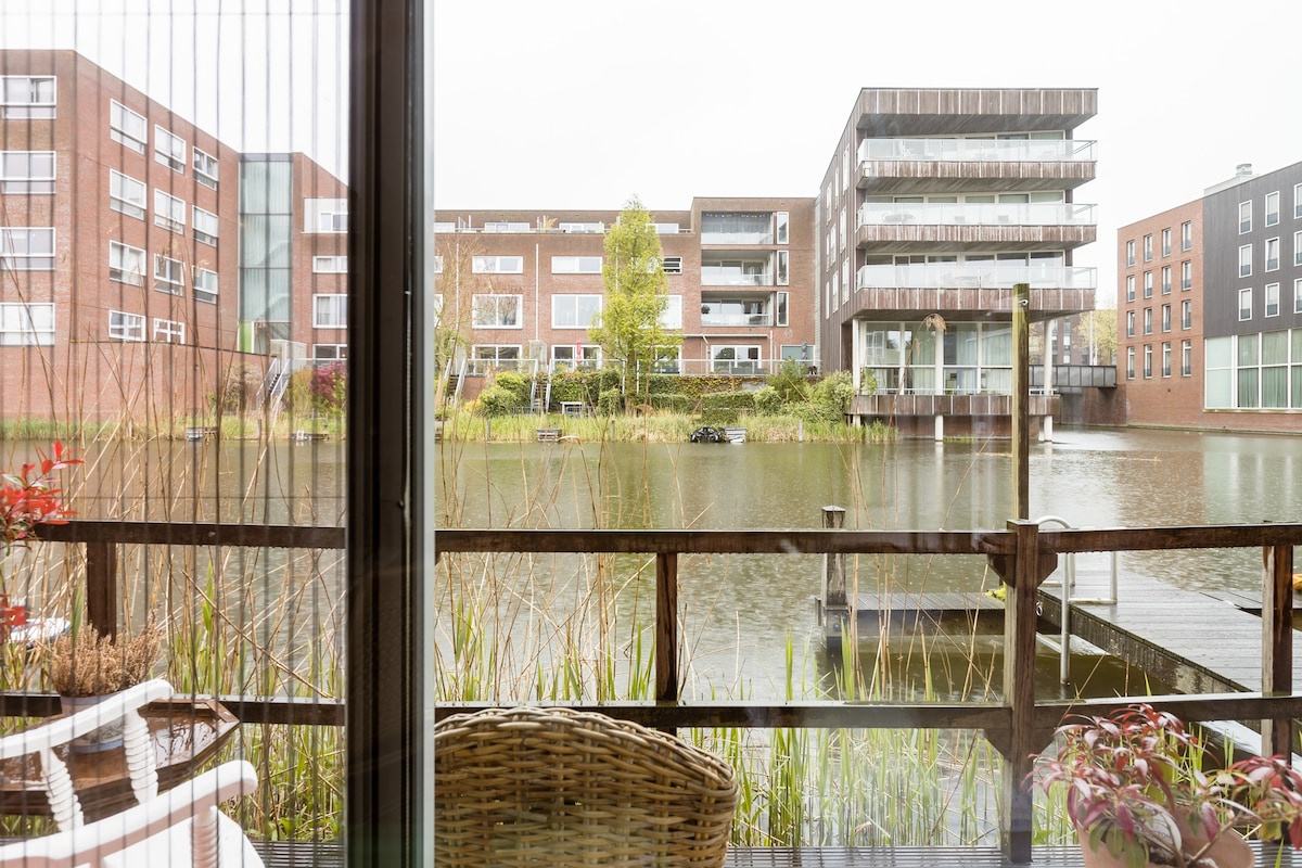 A view from the deck reveals a tranquil water scene with surrounding buildings and a dock. The still water reflects the greenery and architecture, creating a serene atmosphere. Reeds frame the view, adding a natural element to the setting.