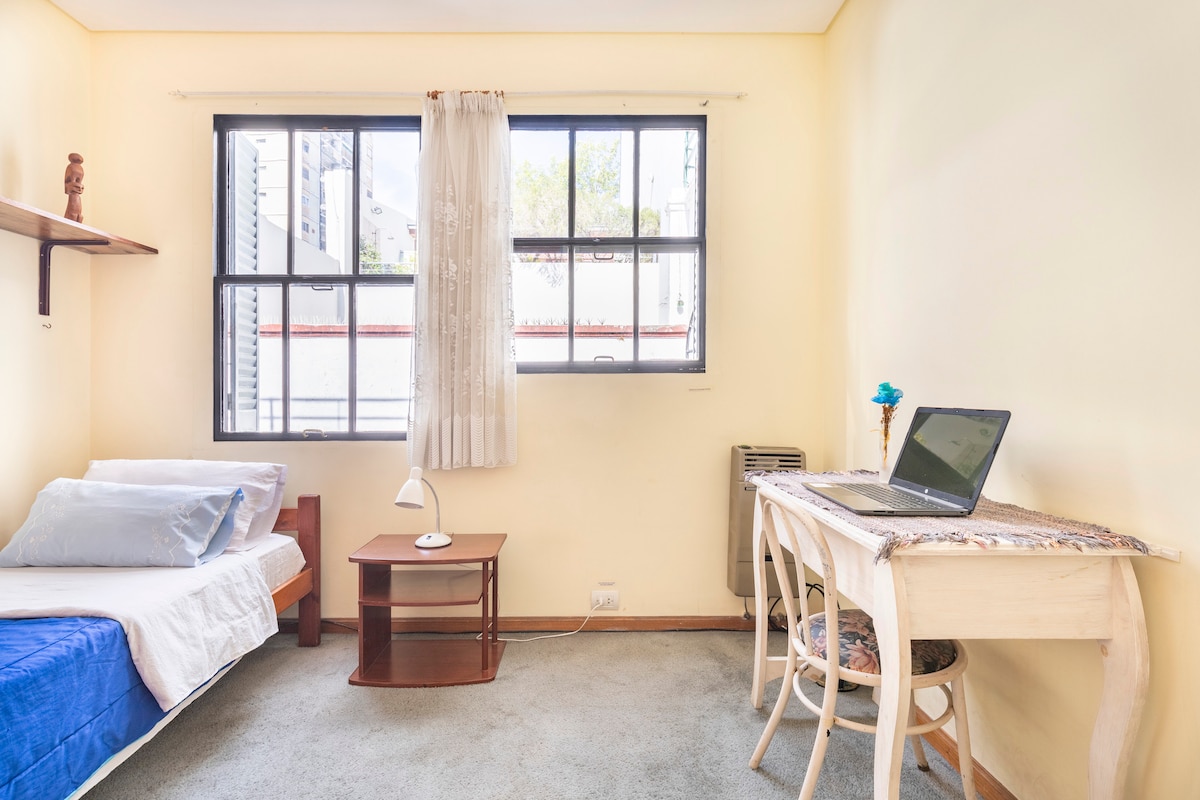 A serene room features a single bed with a blue and white bedding arrangement. Natural light enters through two large windows, providing views of the outdoor space. A small desk with a lamp and a chair enhances the room's functionality, while a cozy carpet adds warmth.
