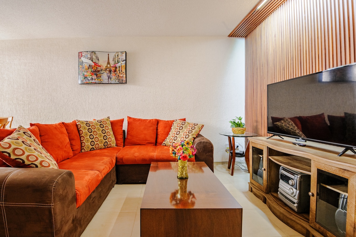 A spacious living area features a large sectional sofa adorned with colorful cushions. A wooden coffee table is positioned in front, accompanied by a flat-screen television mounted against a textured wall. A potted plant adds a touch of greenery, enhancing the inviting layout.