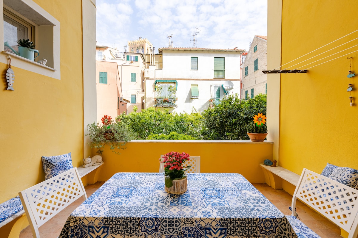 An outdoor terrace is presented, featuring a table adorned with a blue-patterned tablecloth. White chairs surround the table, with potted plants adding a touch of greenery. Yellow walls frame the view of neighboring buildings and clouds above, creating a bright and open atmosphere.
