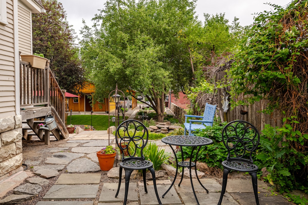 A stone-paved patio area is presented, featuring a round table with two black chairs. Surrounding greenery includes a flowering plant in a terracotta pot and lush grass in the background. A glimpse of a wooden structure and trees adds depth to the outdoor space.