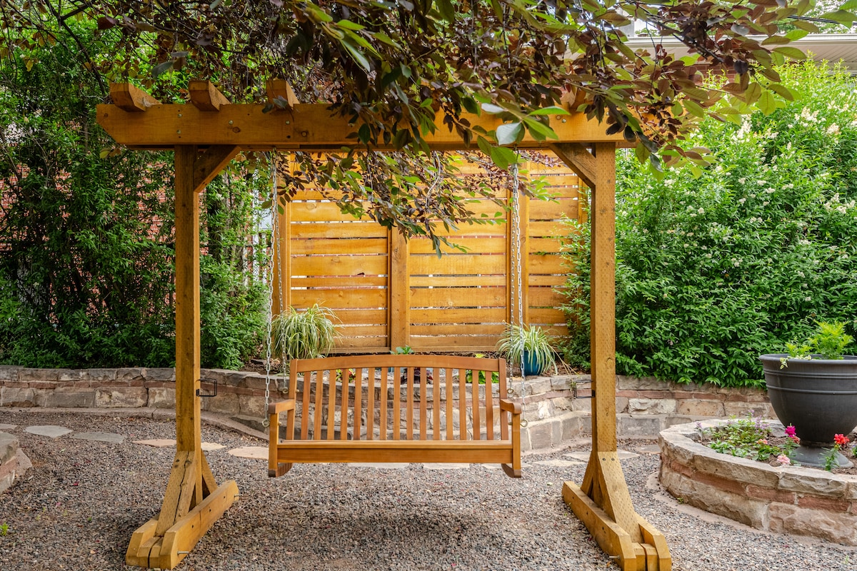 A wooden swing is suspended beneath a canopy of leafy branches, providing a serene spot for relaxation. The swing is framed by a wooden structure, with a backdrop of a privacy fence and lush greenery accentuating the tranquil atmosphere.