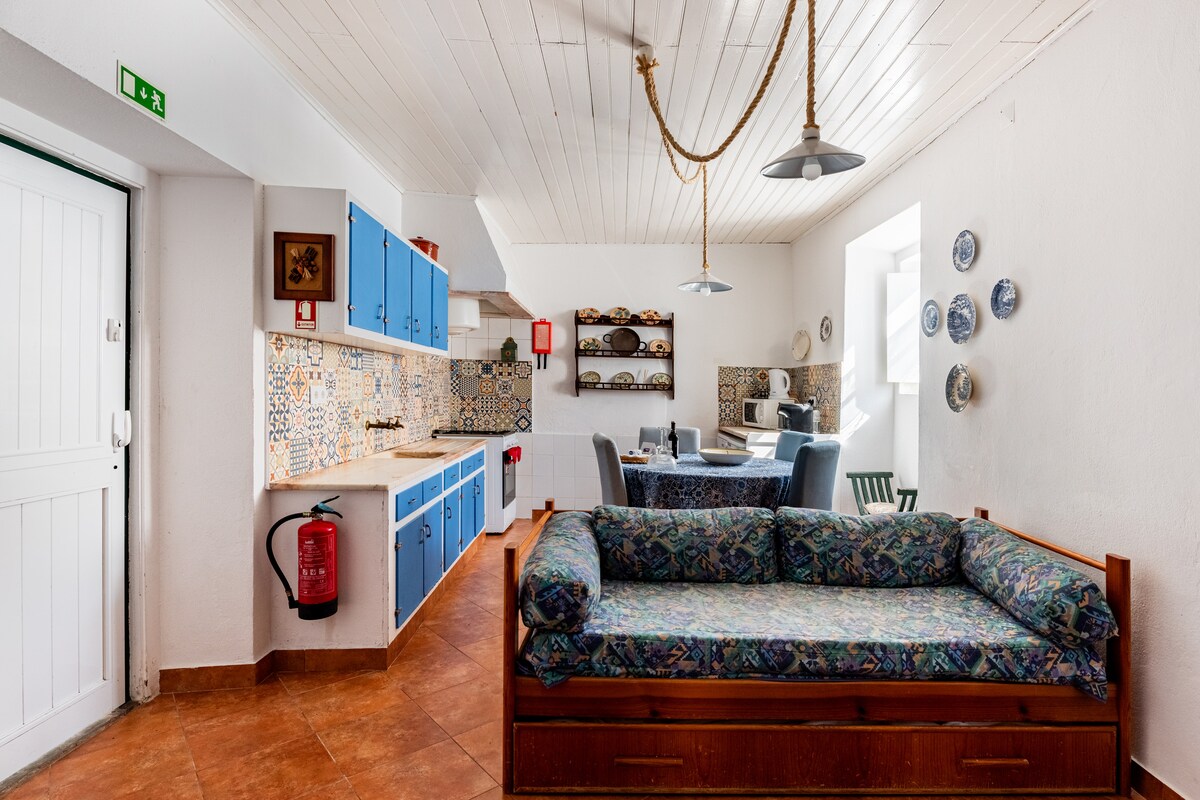 A bright kitchen area features blue cabinetry and patterned backsplash tiles, paired with wooden dining furniture. A cozy seating space with a patterned couch is positioned near a window, which allows for natural light to enhance the atmosphere. Decorative plates are displayed on the wall.