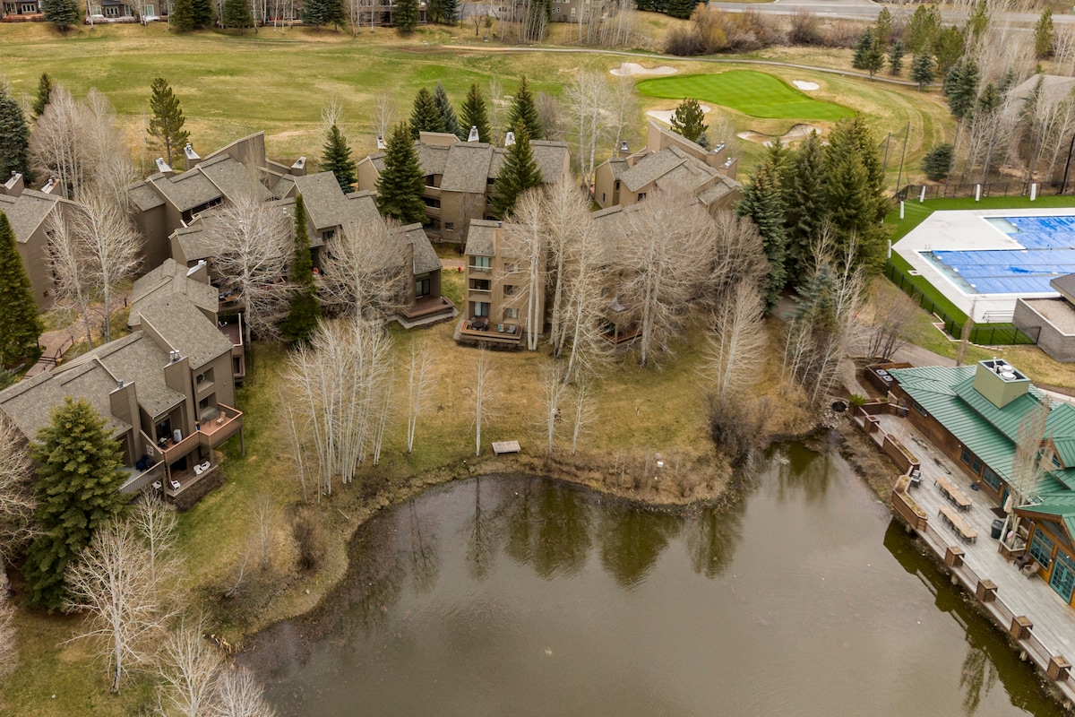 An aerial view captures the scenic layout of a condominium complex surrounded by trees, with a small pond nearby. A golf course can be seen in the background. The property incorporates natural elements, providing a serene and welcoming environment.